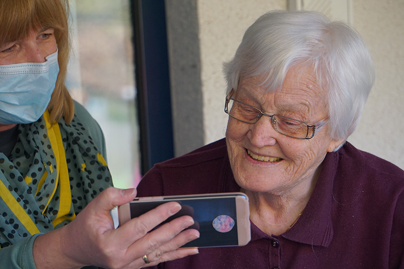 Resident of a nursing home looking at a phone handed to her by a nurse wearing a mask