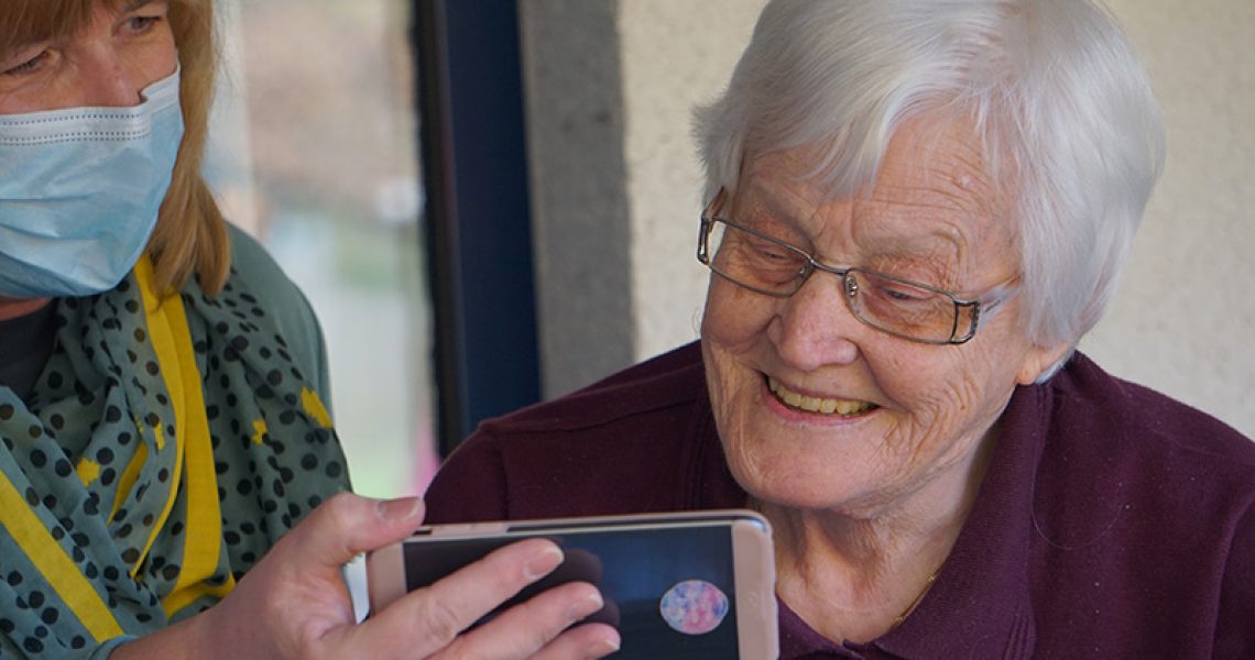 Resident of a nursing home looking at a phone handed to her by a nurse wearing a mask